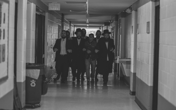 A group of men walking toward the camera in a long, brightly lit school hallway.
