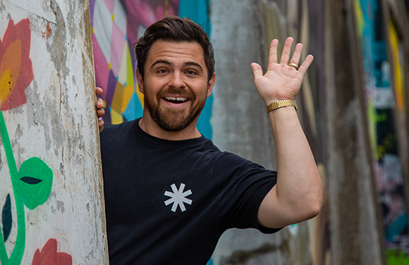 A smiling man with a beard waves his hand while standing next to a colorful graffiti wall.