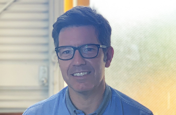 A smiling man with dark hair and glasses wearing a blue shirt.