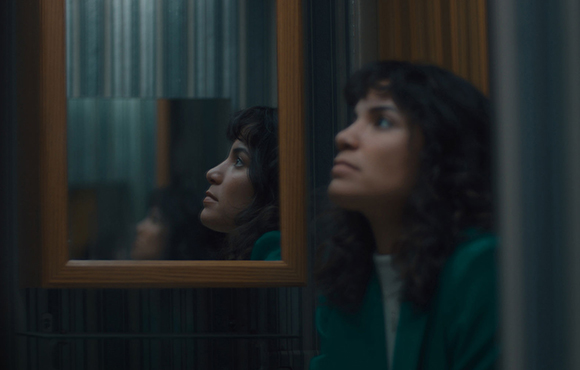 A woman with dark curly hair looks thoughtfully into a framed mirror in a dark, moody setting.