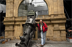 A man in a red jacket stands beside a large, dark vintage steam locomotive in front of a stone building.