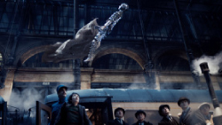 A person in a long cape flies through the air in a dark, arched train station above a steam train.