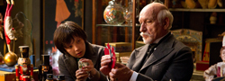 A young boy and an older man with a white mustache examine playing cards in a cluttered shop.