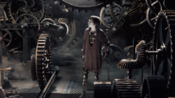 A small figure in a dark robe stands surrounded by large, dark, complex mechanical gears and machinery.