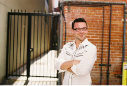 A smiling man with glasses and a white patterned shirt stands with arms crossed in front of a brick wall.