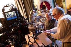 A film crew operates a large camera and monitors on a detailed movie set with various props and equipment.