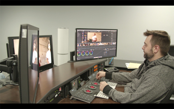 A man sits at a desk with multiple monitors and a video editing control surface.