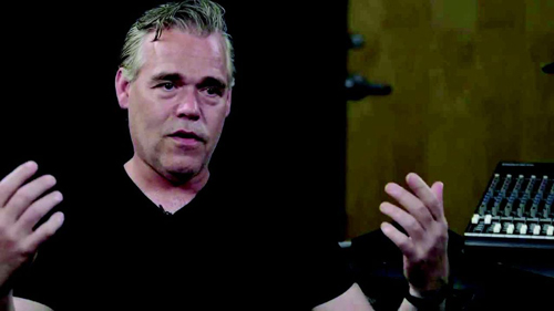 A man in a black shirt gestures with both hands while speaking in a dark studio.