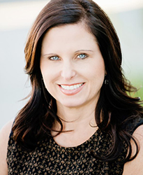 A smiling woman with long dark hair and blue eyes wearing a dark patterned top.