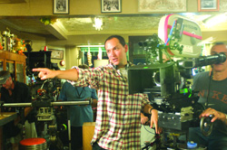 A man in a plaid shirt points toward something while standing next to a large professional film camera.