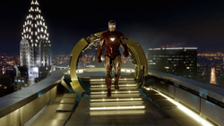 A figure in red and gold armor walks toward the camera on a lit rooftop overlooking a city at night.