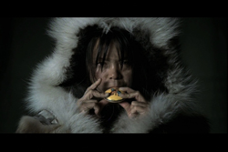 A person wearing a thick, white fur-lined hood looks forward while holding a small, round object near their mouth.