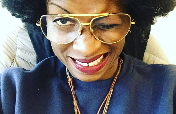 A person with dark curly hair wearing large gold aviator-style glasses and smiling.