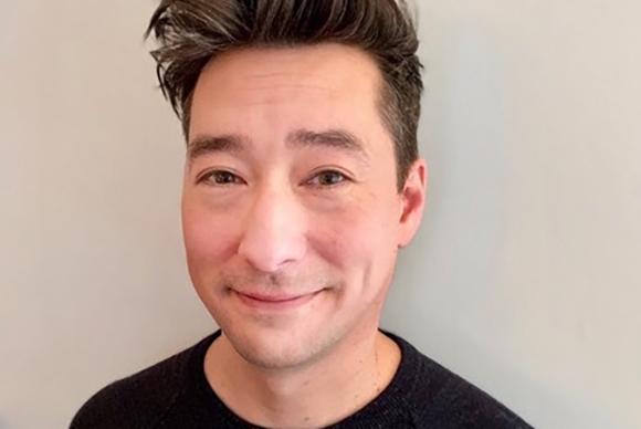 A man with dark hair and a slight smile looking directly at the camera against a plain light background.
