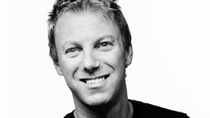 A black and white close-up portrait of a man smiling against a white background.