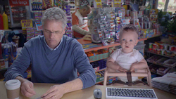 A man in a blue sweater works at a desk next to a baby in a high chair inside a small shop.