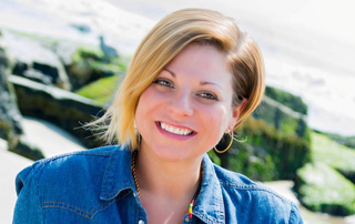 A smiling woman with blonde hair wearing a blue denim shirt in front of a coastal landscape.