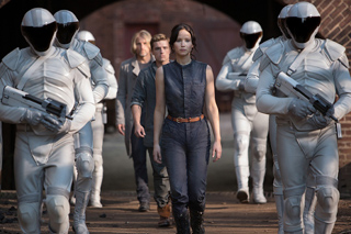 A woman in a dark jumpsuit walks between several figures in white futuristic armor holding weapons.