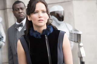A woman with dark hair and a blue fur-trimmed top looks ahead with a worried expression.