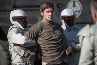 Two people in white futuristic armor hold a man in a brown long-sleeved shirt.