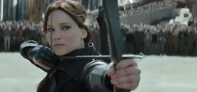 A woman in dark clothing aims a bow and arrow toward the camera with a crowd in the background.