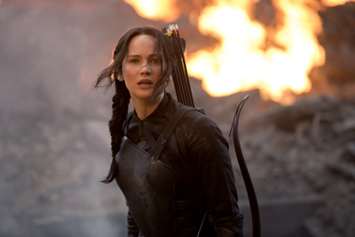 A woman in dark tactical gear with a quiver of arrows looks off-camera against a backdrop of fire and smoke.