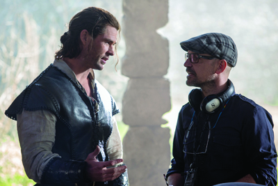 A man in a dark leather vest talks to a man wearing a plaid cap and headphones.