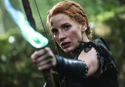 A red-haired woman in dark clothing aims a bow with a glowing blue arrow in a forest.