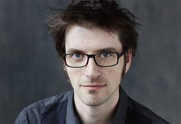A man with dark, messy hair and black-rimmed glasses looking forward against a dark gray background.