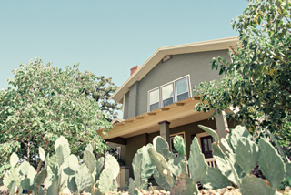 A gray two-story house partially obscured by green trees and prickly pear cacti under a clear sky.