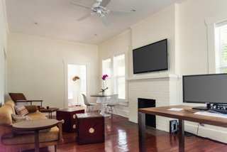 A living room with a white fireplace, wall-mounted TV, tan sofa, and a wooden desk with a computer monitor.