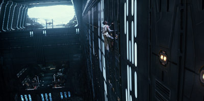 A person in light clothing climbs a tall, dark metallic wall inside a large spacecraft.