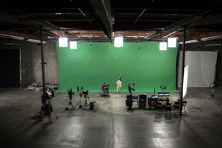 A large green screen set in a dark film studio with cameras, lights, and crew members.