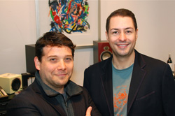 Two men smiling, one in a dark jacket and one in a blazer over a teal t-shirt.