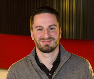 A man with dark hair and a beard smiling, wearing a grey zip-up sweater over a black shirt.
