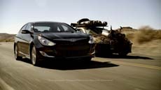 A black sedan drives down a desert highway with a modified off-road vehicle following closely behind.