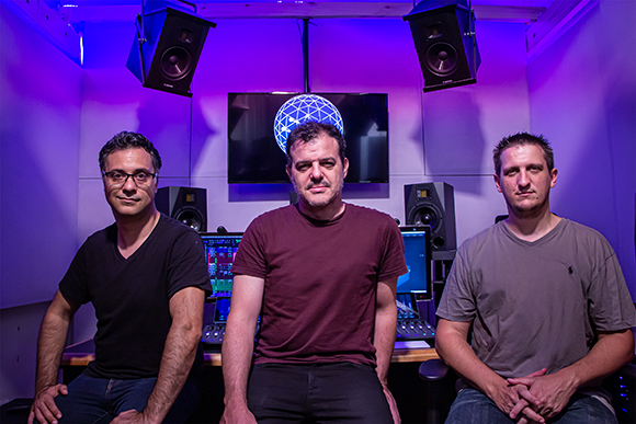 Three men standing in a recording studio with speakers and monitors in the background.