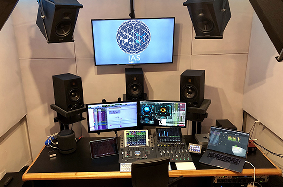 A music studio desk with multiple monitors, studio monitors, a laptop, and audio mixing controllers.