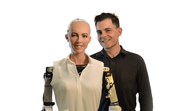 A man in a black shirt smiling next to a humanoid robot wearing a white blouse.
