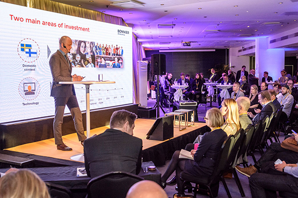 A man stands at a podium presenting a slide titled Two main areas of investment to an audience in a conference room.