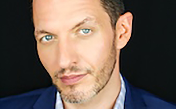 Close-up portrait of a man with light blue eyes and a short beard against a black background.