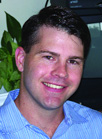 A man with short dark hair smiling while wearing a light blue striped button-down shirt.