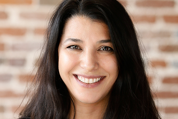 A smiling woman with long dark hair looking directly at the camera against a brick background.