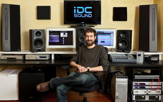 A man sits in a chair in a studio filled with large speakers, monitors, and mixing equipment.