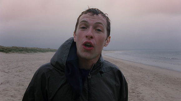 A man in a dark hooded jacket stands on a sandy beach under a cloudy sky.