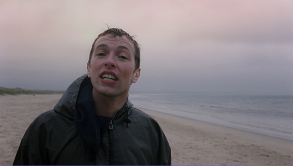 A man in a dark hooded jacket stands on a sandy beach with the ocean in the background under a gray sky.