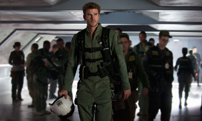 A man in a green flight suit and tactical gear walks forward through a hangar filled with other personnel.