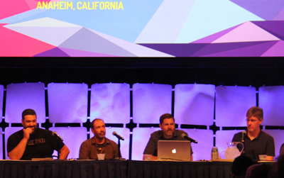 Four men sit at a table on a stage. A large screen behind them displays "ANAHEIM, CALIFORNIA" and a geometric pattern. The me