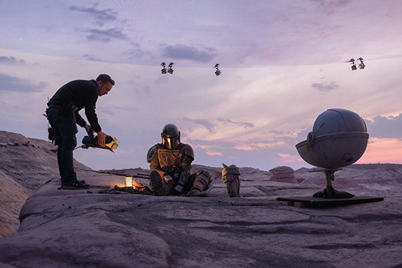 A man stands by a person in Mandalorian armor sitting by a campfire in a desert at dusk.