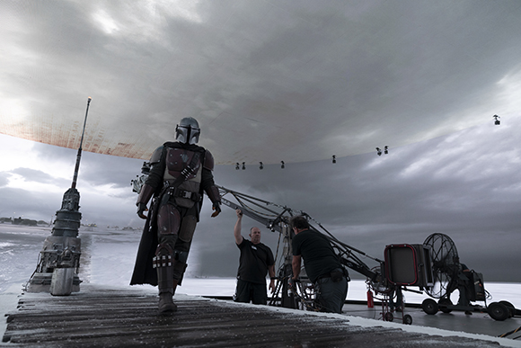 A person in Mandalorian armor stands on a set facing a crew and a large overhead lighting dome.